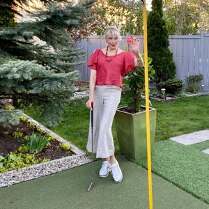 Model wears beige linen wide leg cropped Jill Pant and coral linen Flutter top. She holds a golf ball having just made a whole in one on her backyard putting green. She is 5'7" tall and wears XS