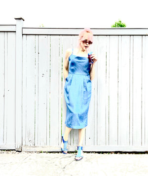 Model leans against a fence drinking from a straw. She wears a blue denim Tencel dress with pleats and large pockets. This version has single straps. She is 5'7" tall and wears size S