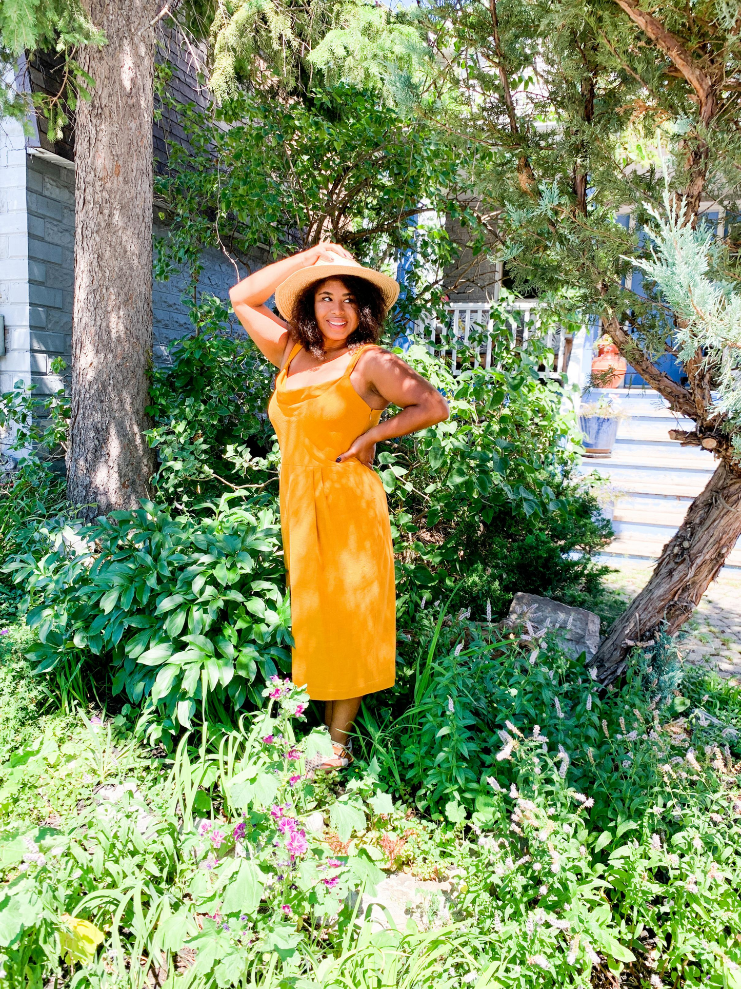 Model stands in garden wearing saffron yellow retro style Lindon dress with pleats at waist and large pockets and bias cut bodice. She is 5'3" tall and wears XL