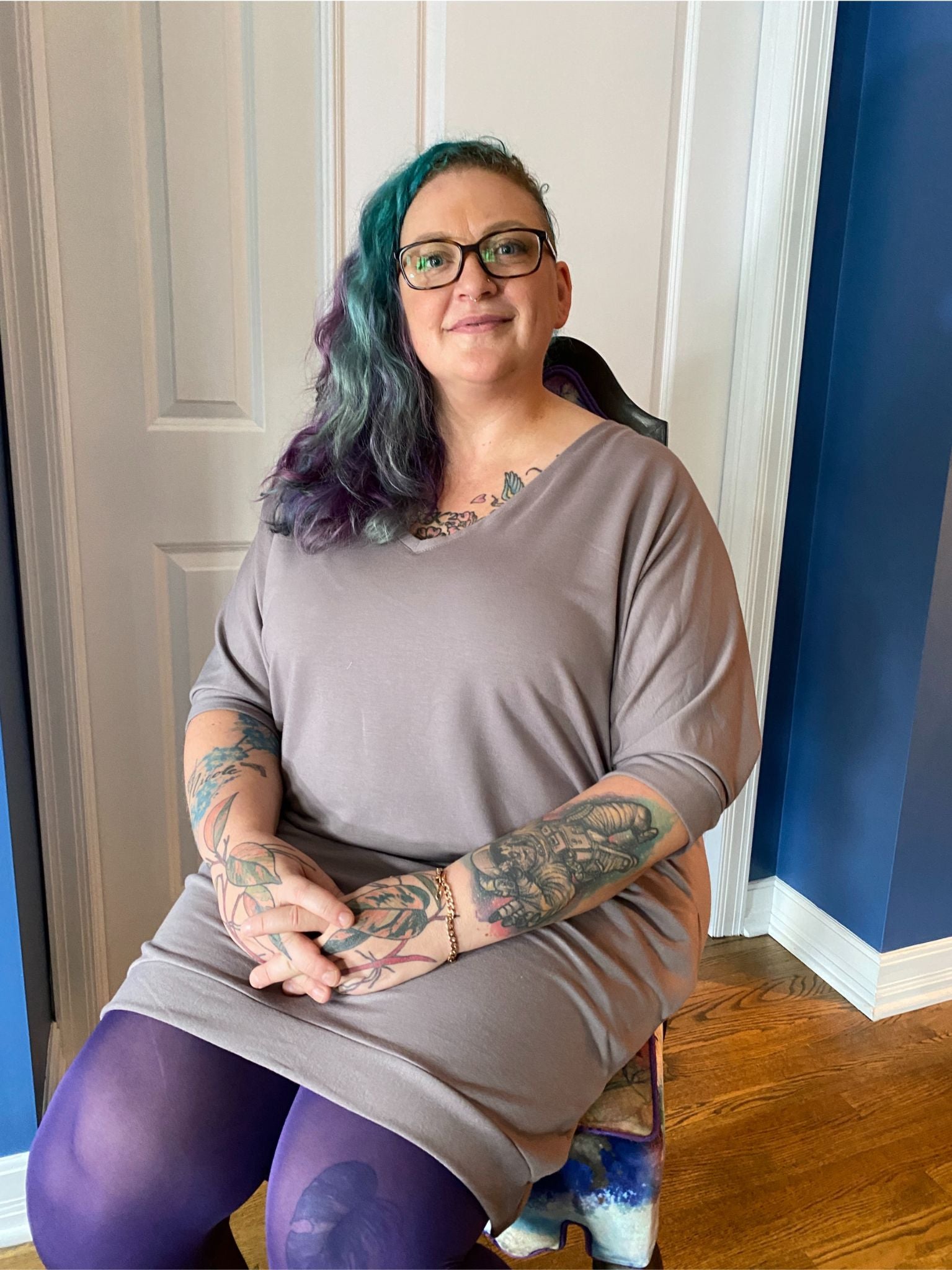 Kristin wears a 2X Ava Sweater dress in mink bamboo/cotton French terry with purple tights to match her purple and turquoise hair. She sits on a chair.