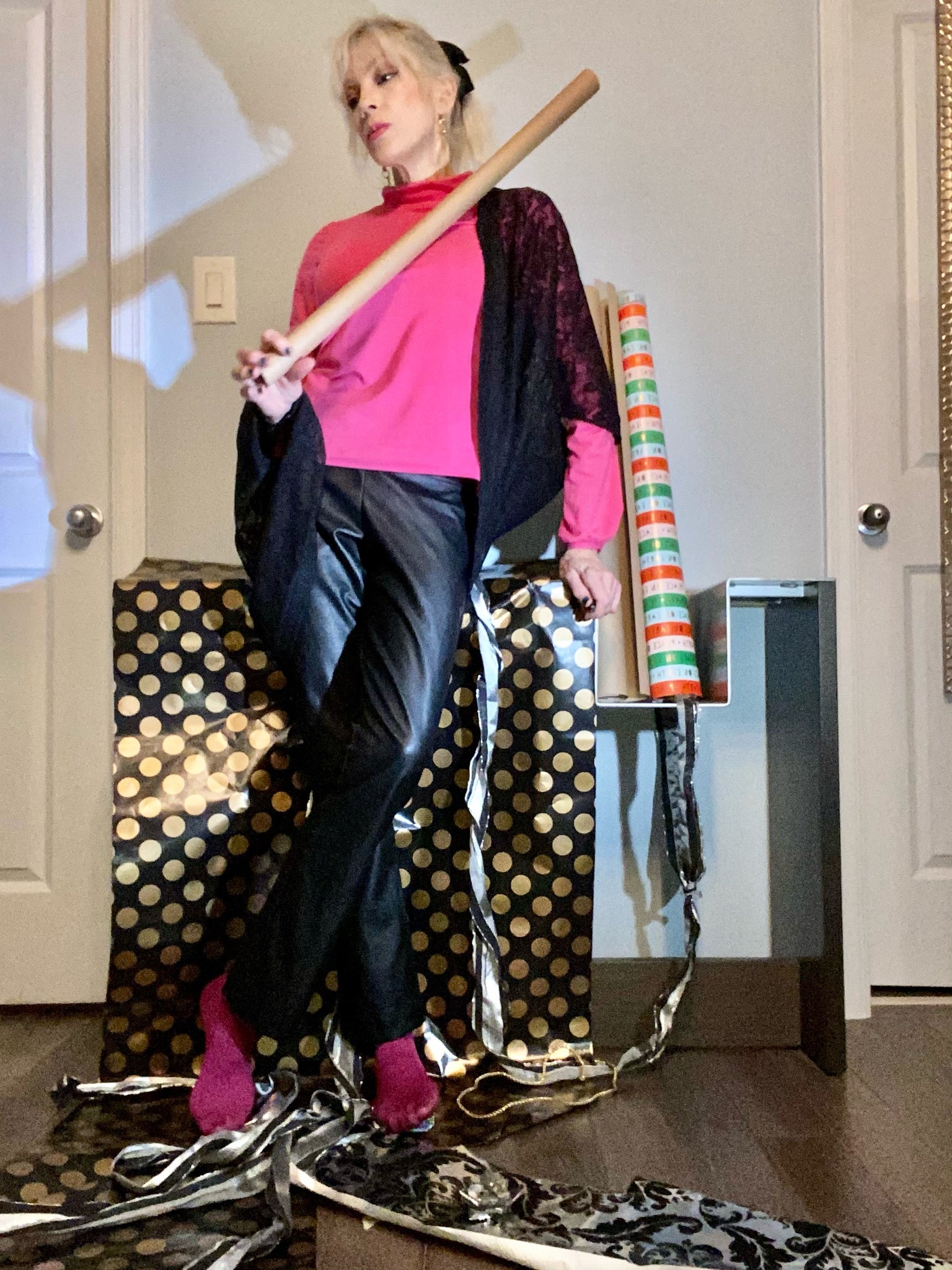 Judy wears a pink turtleneck with short 60s-inspired collar and gathered modified bishop sleeves. She paired it with black leatherette Kissin' pants and a black lace Pippa shrug. She stands in a mess of wrapping paper and holds an empty wrapping paper roll